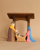 The Ostheimer Nativity Scene showcases a minimalist stable design that encourages imaginative play. It includes figures such as a character in a brown and yellow cloak, another dressed in a blue and pink robe, and an infant lying in a manger, all set against a gentle beige background.