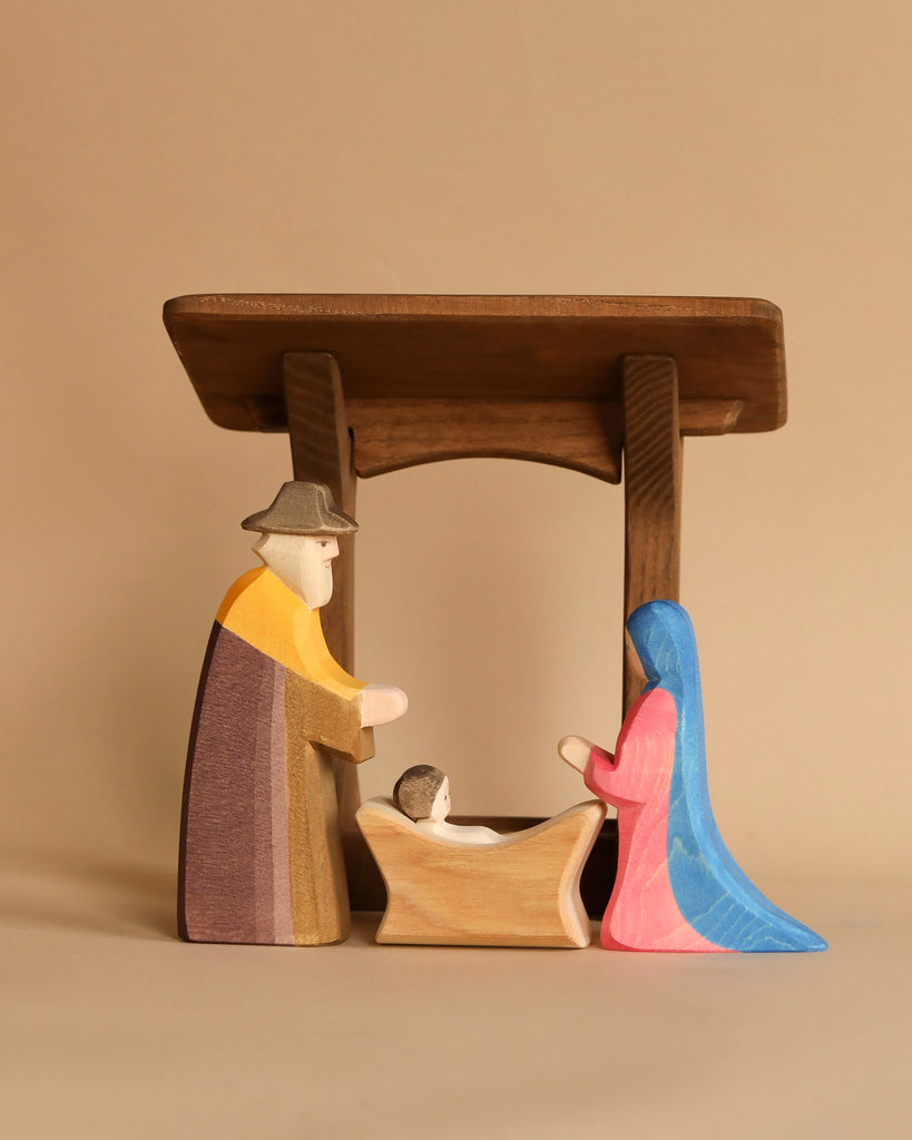 Ostheimer Nativity Scene - Extended: A wooden nativity scene crafted from sustainable materials, featuring a simple stable and three figures—a child in a manger, an adult in a hat on the left, and an adult with a blue cloak on the right. Designed for imaginative play, this set is complemented by a warm, neutral-toned background.