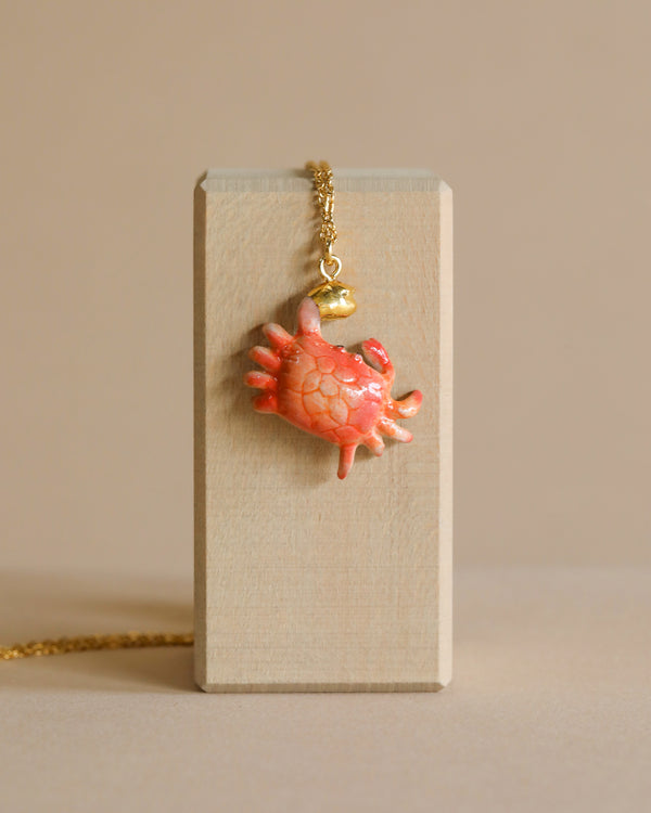 A delicate, anatomical heart pendant crafted from porcelain hangs from a Crab "Golden Grip" Necklace, displayed on a vertical, rectangular wooden block with a beige background.