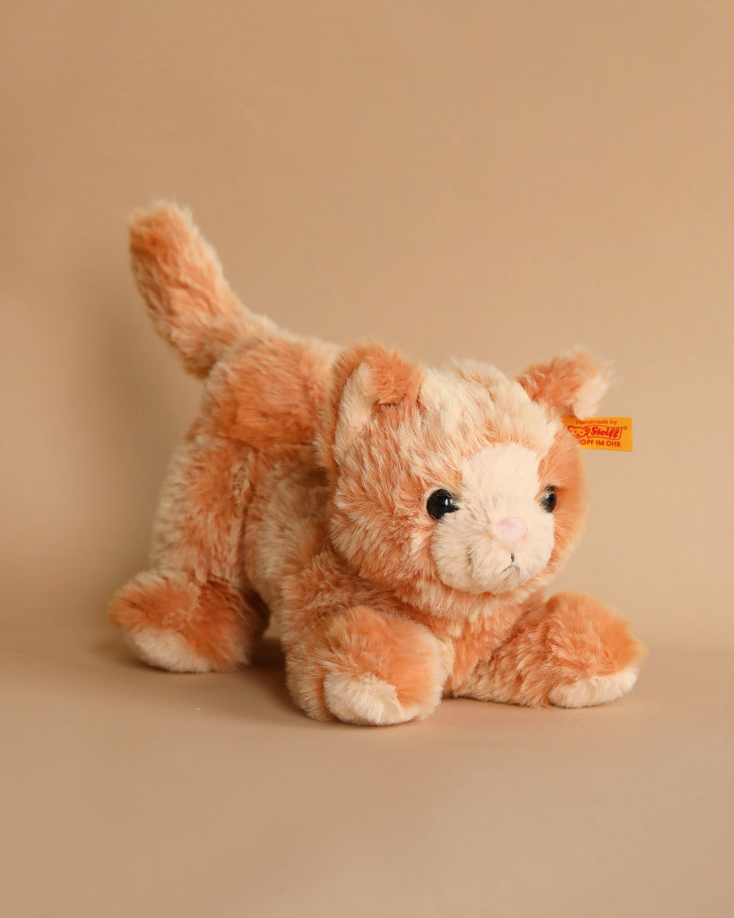 A Cassie Cat Plush Toy with orange and white fur sits on a neutral beige background. This huggable companion features a label attached to its ear, inviting you to embrace its soft charm.