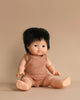 A Minikane Clothed Standing Doll (14.5") - Jude-Léo with short, black hair sits on a light brown background. The anatomically correct doll is dressed in a sleeveless, light brown ribbed outfit and has a neutral expression. Its legs are extended forward, and its arms rest by its sides. This doll features a subtle natural vanilla scent.