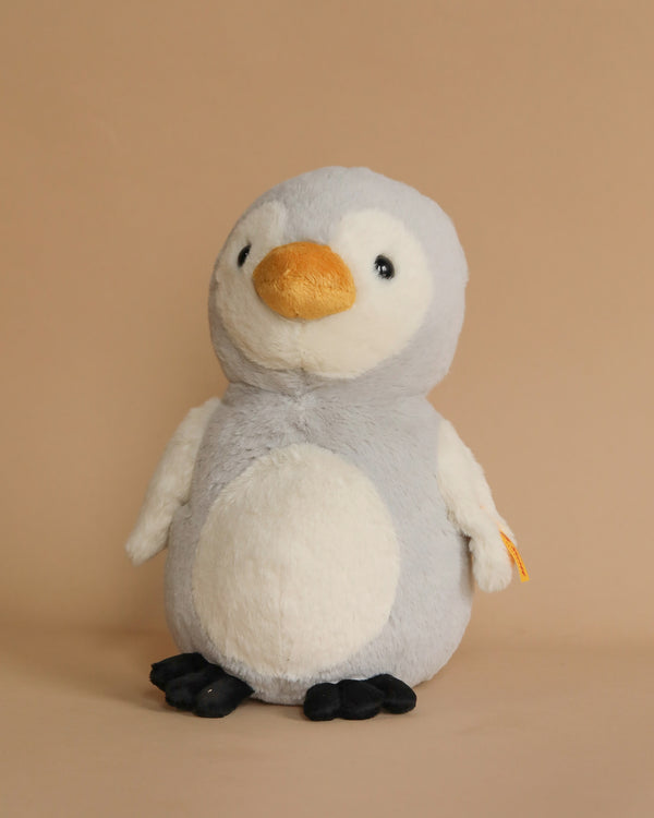 The Steiff Penguin Stuffed Plush Animal, featuring a soft grey body, white belly, and orange beak, stands on a neutral background, ready to be your cuddly adventure playmate.