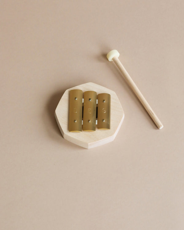 A small wooden toddler instrument, the Auris Glockenspiel 3 tones d-e-g features an octagonal shape with three gold metal bars and a light-colored mallet with a white tip, all placed on a beige surface.