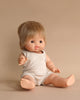 A Minikane Clothed Standing Doll (14.5") - Archie with short, light brown hair is sitting on a beige surface. It wears a sleeveless, ribbed, off-white romper and has a neutral expression. Made in Spain, the anatomically correct doll sits against a matching beige background, giving the image a soft, monochromatic look.