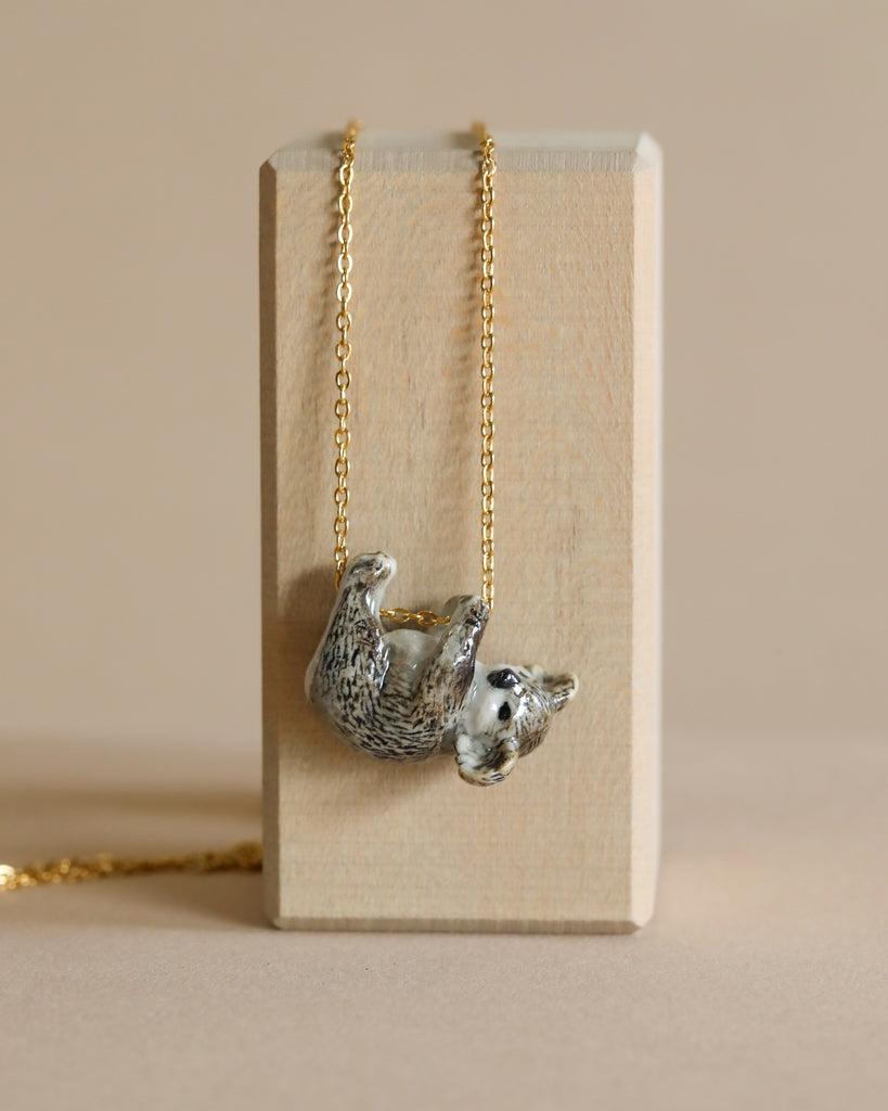 A delicate gold plated chain necklace featuring a small, intricately detailed koala pendant hanging upside down. The koala is positioned on a simple, rectangular wooden display against a plain beige background. The design showcases the koala's textured fur and relaxed pose, embodying hand painted jewelry craftsmanship.