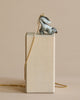 A pendant necklace featuring a silver skunk, suspended on a thin 24k gold plated steel chain, draped over a tall beige box against a neutral background.