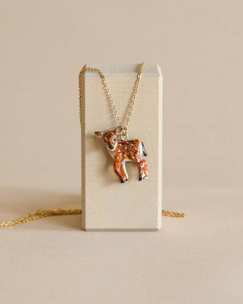 A Fawn Necklace, hand painted and hanging on a 24k gold plated chain, displayed against a beige block on a light background.