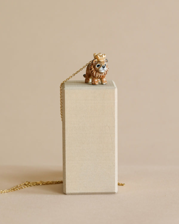 The image shows a whimsical Lion King Necklace with a crown, positioned on top of a rectangular beige block. A delicate gold chain is attached to the necklace, draping gently over the side of the block. The high-quality porcelain pendant is hand-painted in detail, set against a matching beige background.