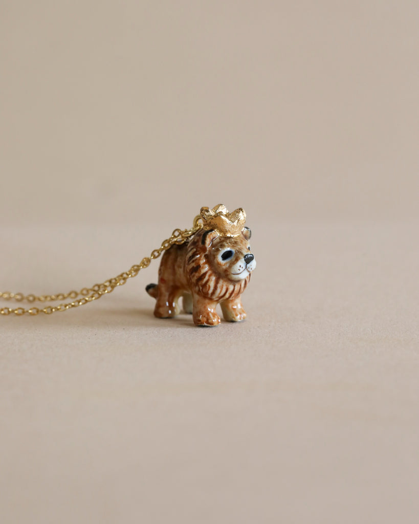 A small, adorable Lion King Necklace, hand painted in detail, with a gold crown atop its head is attached to a gold chain, set against a light beige background. The lion has a cute, cartoon-like face with large eyes, adding a whimsical touch to this high-quality porcelain piece.