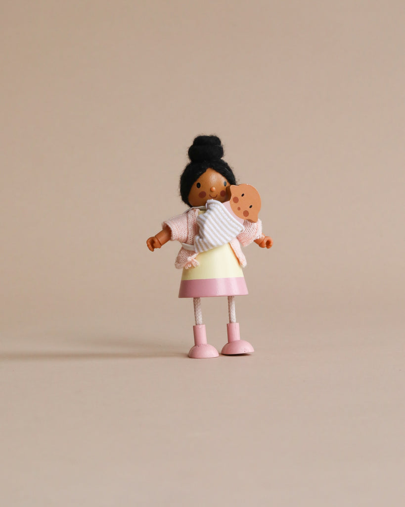 A small doll with dark hair styled in a bun, wearing a white and pink dress made of knitted and jersey fabrics, holding Mrs. Forrester and the Baby against a plain beige background.