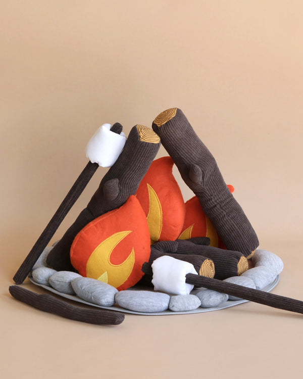 A Campout Campfire & S'mores set resembling a cozy campfire scene. The set includes fabric logs, stone-like pillows, faux felt flames, and marshmallows on sticks for pretend s’mores. The pieces are arranged to mimic a plush campfire on a neutral beige background.