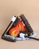 A Campout Campfire & S'mores set resembling a cozy campfire scene. The set includes fabric logs, stone-like pillows, faux felt flames, and marshmallows on sticks for pretend s’mores. The pieces are arranged to mimic a plush campfire on a neutral beige background.