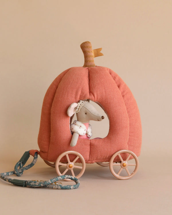 A small stuffed mouse toy from the royal mice family sits inside a soft, fabric fairytale carriage with a handle for pulling. The Maileg Pumpkin Carriage is orange with a stem on top and four wheels at the bottom. The mouse, dressed in a white outfit, peeks through a window in the carriage.