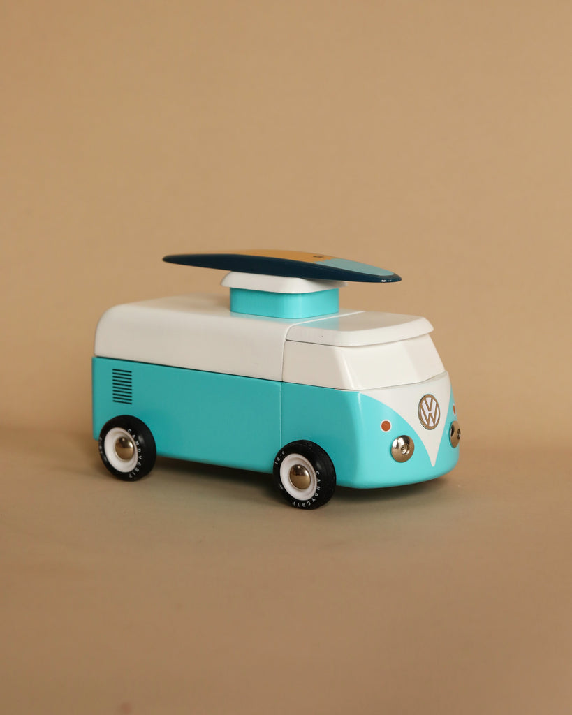 The VW Beach Bus Ocean toy model, a vintage Volkswagen Type 2 van with a surfboard on top, painted in teal and white, is positioned against a plain beige background.