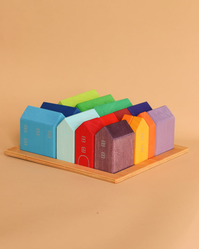 Grimm's Houses, a collection of vibrant, hand-painted wooden block houses arranged on a wooden tray, evoke the imagination of a city planner's dream with their lively colors including blue, green, red, purple, orange, and yellow against a simple beige backdrop.