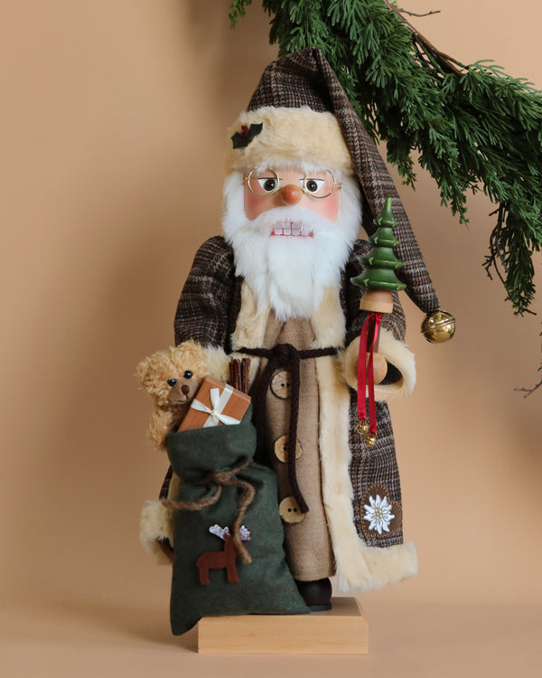 A festive Santa Claus figurine, dressed in a brown checkered coat and hat, holds a small Christmas tree and a sack filled with gifts, including a teddy bear. Greenery is elegantly draped above, set against a neutral background. The scene is further enhanced by the Christian Ulbricht Collectible Wooden Nutcracker for an extra touch of holiday charm.