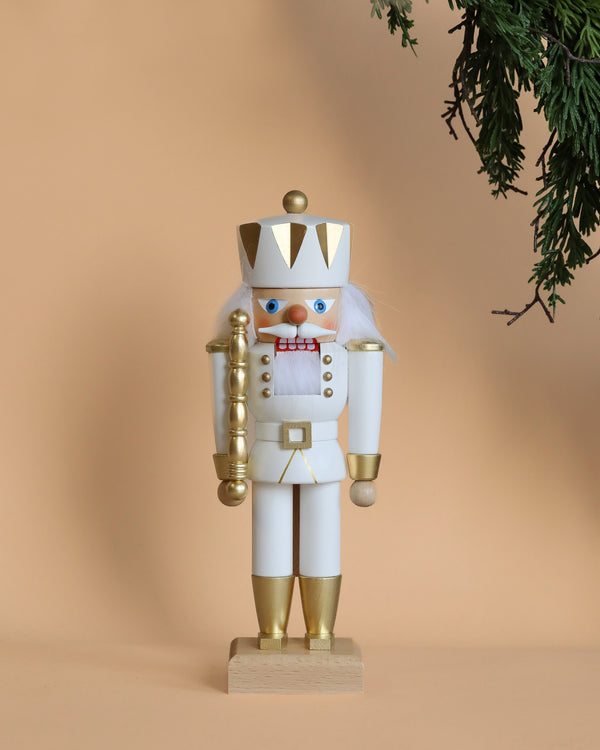 A Christian Ulbricht Collectible Wooden Nutcracker - King (White) stands upright against a beige background. It holds a gold staff and features a red and white checkered bow tie, while pine branches frame the top right corner.