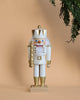 A Christian Ulbricht Collectible Wooden Nutcracker - King (White) stands upright against a beige background. It holds a gold staff and features a red and white checkered bow tie, while pine branches frame the top right corner.