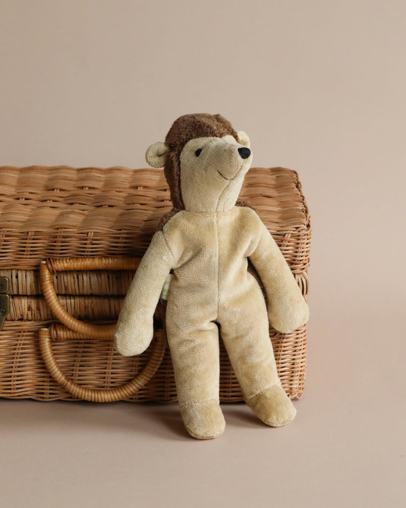 A plush Senger Naturwelt stuffed animal - Hedgehog handmade in Germany in a soft beige color, seated against a woven wicker basket on a plain beige background. The stuffed animal appears soft with a content facial expression.