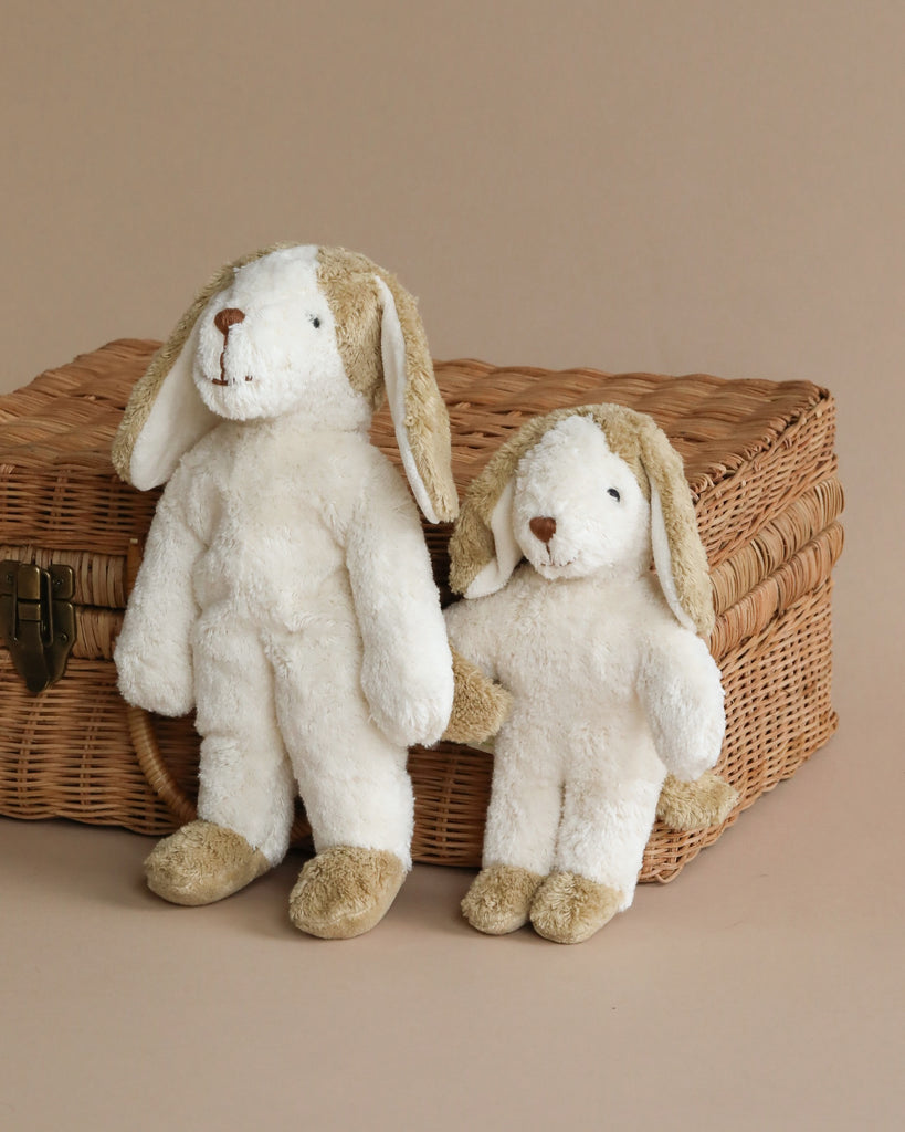 Two plush rabbit toys seated against a Senger Naturwelt Stuffed Animal - Dog basket on a beige background. One toy is upright, and the other is leaning slightly, both looking forwards.