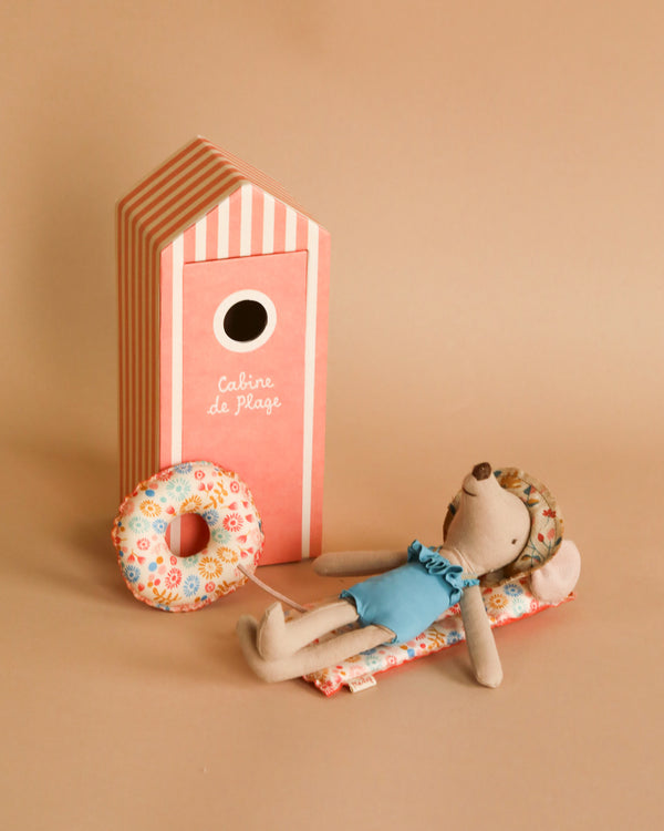 A small toy mouse in a blue bathing suit and floral bonnet lies on a patterned mat with a floral inflatable ring nearby. Behind the mouse is a pink and white striped beach house with the text "Cabine de Plage" on it, all set against a beige background. This scene features the Maileg Beach Mom Set.