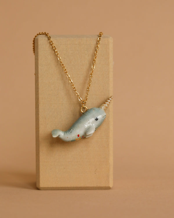 A gold chain necklace displayed on a wooden stand features the Narwhal Necklace with a white tusk and black eyes against a plain beige background.