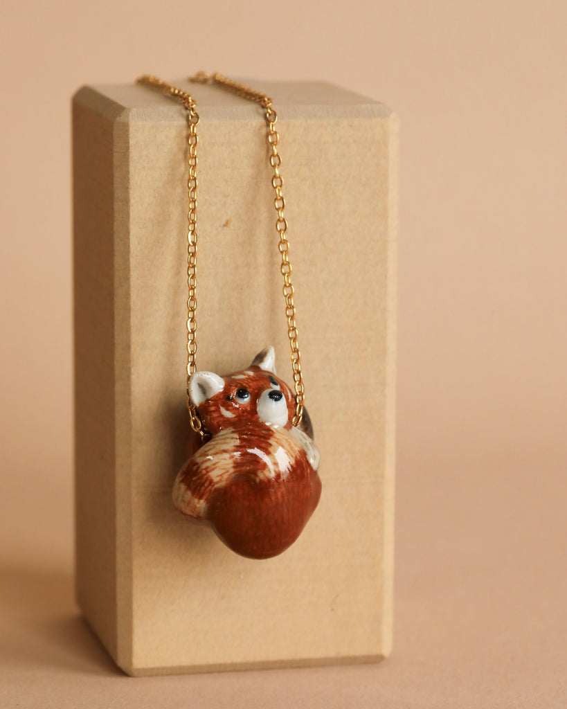 A Red Panda Necklace with a 24k gold plated chain featuring a ceramic pendant shaped like a red panda's head. The heirloom quality necklace is displayed against a beige background, hanging from a light wooden block.