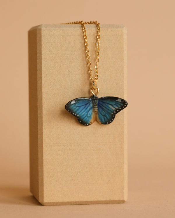 A delicate Blue Butterfly Necklace featuring a blue butterfly pendant dangles against a light wooden display block. The 24k gold plated steel chain adds an elegant touch to the handcrafted porcelain pendant, showcasing intricate details on the butterfly's wings set against a beige background.