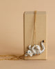 A delicate 24k gold plated steel chain necklace with a hand-painted porcelain white tiger pendant draped over a rectangular beige box. The playful tiger lies on its back with blue eyes, black stripes, and a small orange detail near its tail. The background is a neutral tone. Product Name: Gold Tiger Necklace