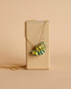 The Chameleon Necklace, with a colorful, hand-painted pendant shaped like a fish, is displayed on a light wooden block against a beige background. The 24k gold plated steel chain complements the fish pendant, which features vibrant yellow, blue, and green stripes with small red and white dotted accents.