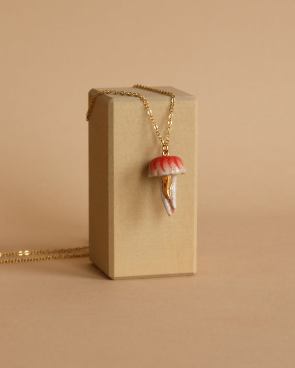 A Jellyfish Necklace with a pendant shaped like a red and white mushroom hangs over a rectangular beige stand against a matching beige background. This 24k gold-plated piece boasts heirloom quality, with the mushroom pendant showcasing detailed features, including a textured cap and stem.