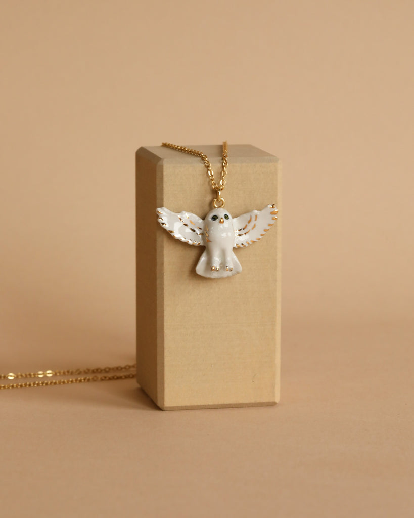 A stunning Gold and White Snowy Owl Necklace, 24k gold plated and hand-painted, is elegantly displayed hanging on a beige rectangular stand against a matching background, highlighting its exquisite craftsmanship.