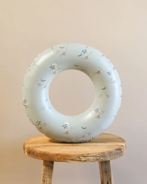 An Inflatable Junior Swim Ring - Dinosaurs made from durable PVC, with patterns of dinosaurs and birds, is propped up on a simple wooden stool against a plain beige background. Perfect for kids aged 3-6 years.