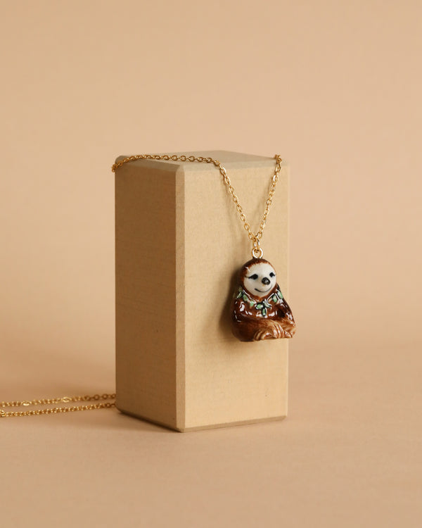 A Happy Sloth Necklace is displayed hanging on a beige rectangular box against a matching beige background. The pendant dangles from a 24k gold plated steel chain, showcasing intricate hand-painted details.