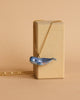 A Blue Whale Necklace with a hand-painted blue whale pendant is draped over a rectangular, beige-colored block. The background is a matching beige, creating a minimalist and cohesive composition.