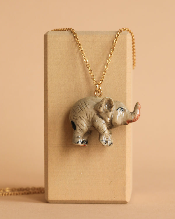 A Baby Elephant Necklace is displayed against a beige background. The pendant is intricately detailed with a textured surface and slight color variations, adding to its realistic portrayal, showcasing heirloom quality craftsmanship.