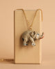 A Baby Elephant Necklace is displayed against a beige background. The pendant is intricately detailed with a textured surface and slight color variations, adding to its realistic portrayal, showcasing heirloom quality craftsmanship.