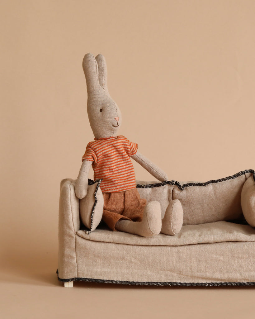 A Maileg Rabbit Size 1 - Classic T-shirt & Shorts with beige fur, wearing an orange and white striped shirt and brown shorts, is sitting on a matching beige couch made from natural linen. The background is a plain light brown color, complementing the neutral tones of the rabbit and the dollhouse furniture.