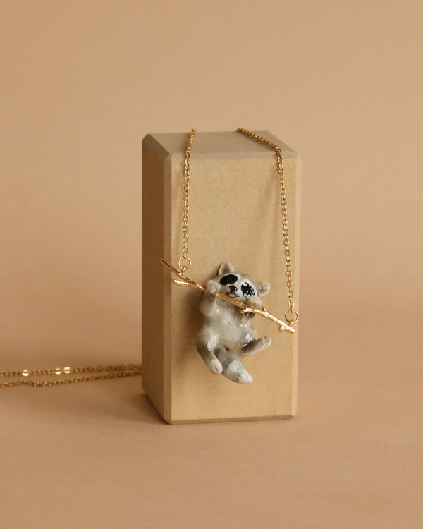 A handcrafted pendant featuring a clay raccoon with a gold chain is displayed on a tan rectangular block against a beige background. Holding a thin gold branch in its arms, the playful and charming raccoon adds an endearing touch to this unique Rascal Raccoon Necklace.