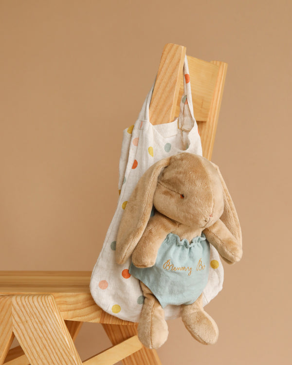 A plush Maileg Bunny Bob, dressed in a blue outfit, is hanging from a vibrant polka dot bag. The beige tote, adorned with colorful spots, is casually draped over the backrest of a wooden chair set against a plain beige background.