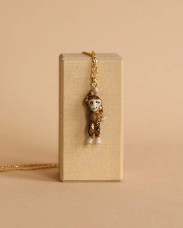 This Sasquatch Necklace features a small figurine pendant on a gold chain. The pendant, crafted in handcrafted jewelry fashion, showcases a detailed brown and white creature with a facial expression, hanging from a wooden block against a beige background. Truly limited-edition fashion.