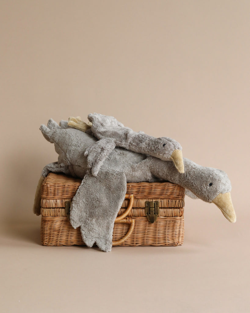 A Senger Naturwelt Cuddly Animal - Grey Goose plush toy resembling a goose, draped over a closed wicker picnic basket, set against a plain beige background.