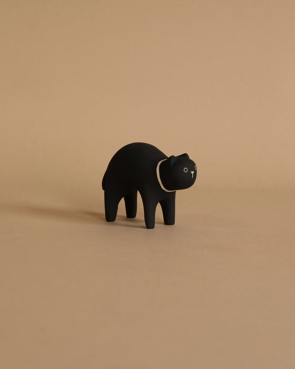 A small, hand-painted black figurine of a cat stands on a beige background. The cat has a simple and minimalist design with white eyes and a small white snout.