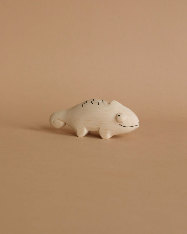 A small, hand-carved Albizia wood handmade tiny wooden animals - chameleon figurine, crafted by skilled artisans in Bali, features simple black line details for eyes, a mouth, and patterned markings on its back. Set against a peach-colored background, the light beige chameleon has a charming curved tail.