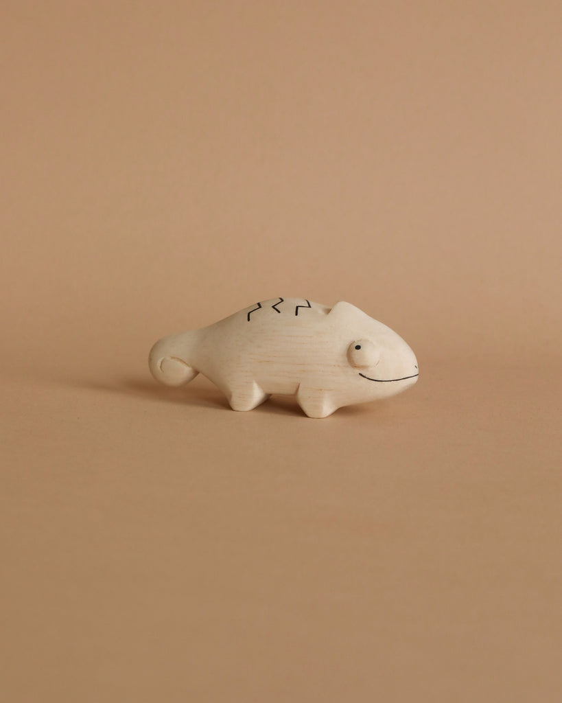 A small, hand-carved Albizia wood handmade tiny wooden animals - chameleon figurine, crafted by skilled artisans in Bali, features simple black line details for eyes, a mouth, and patterned markings on its back. Set against a peach-colored background, the light beige chameleon has a charming curved tail.