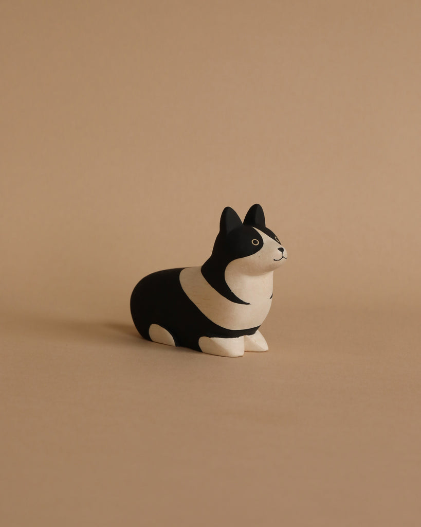 A small, black and white **corgi dog figurine** with a minimalist design and rounded body sits on a plain beige surface, against a matching beige background. Hand-carved by Albizia wood artisans, the corgi has a curious expression and pointed ears.