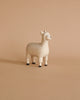 A small, white wooden figurine of a llama, **Handmade Tiny Wooden Farm Animals - Alpaca**, hand carved from Albizia wood by artisans in Bali, stands against a plain, beige background. The figurine has black-tipped feet and a simple, cute expression on its face.