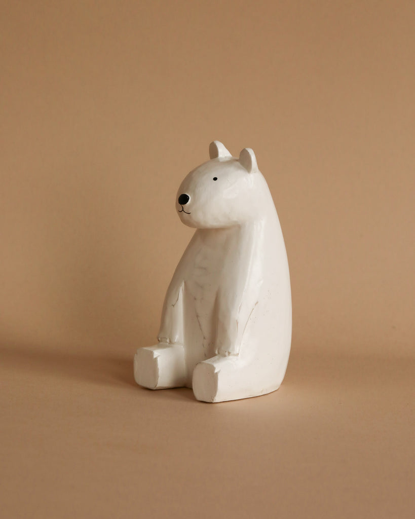 A small, white ceramic figurine of a bear sits upright against a light brown background. The Handmade Tiny Wooden Polar Bears have simple black eyes and a small nose, with no other details or colors. The minimalistic design resembles handcrafted wooden animals, giving it a cute, playful appearance.