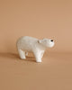 A small, white, ceramic figurine of a bear with a simplistic, minimalist design stands on a light brown surface. The backdrop is a matching light brown, creating a cohesive, earthy aesthetic. The Handmade Tiny Wooden Polar Bears have a gentle expression with minimal black nose and eyes, reminiscent of handcrafted wooden animals.