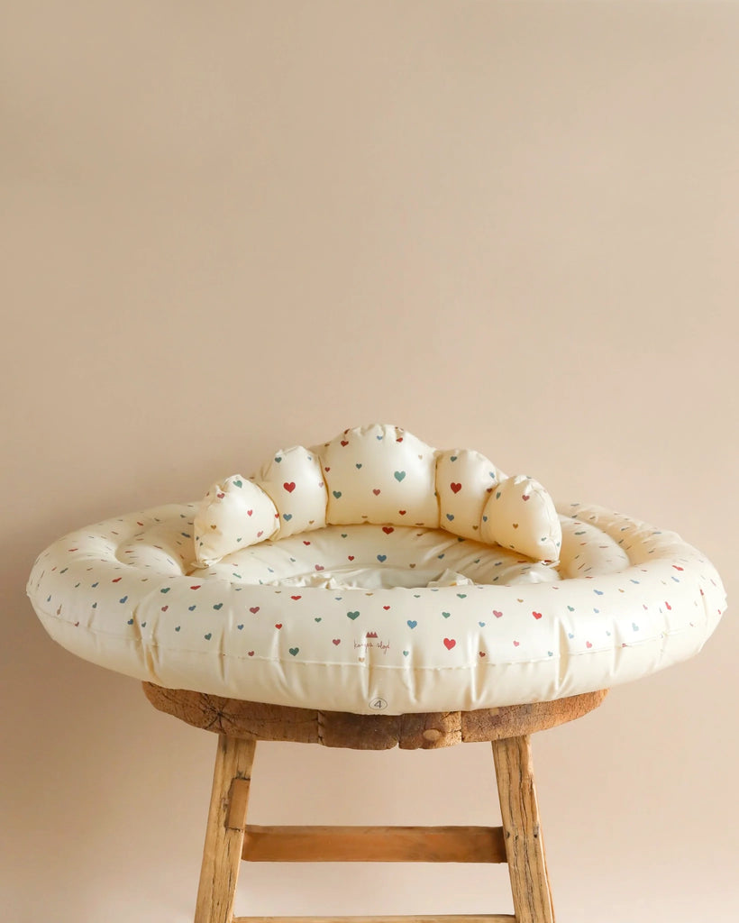 A round, cream-colored Inflatable Baby Swim Ring - Multi Hearts is placed on a wooden stool. The background is a solid, light beige color. The cozy and secure swim ring for infants features a padded backrest and durable PVC construction that complies with EN 13138-3:2021 standards.